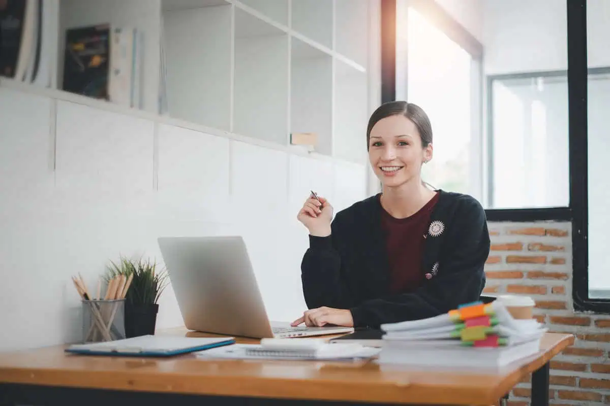 Smiling professional woman sitting at a desk with a laptop, documents, and office supplies in a bright and modern workspace