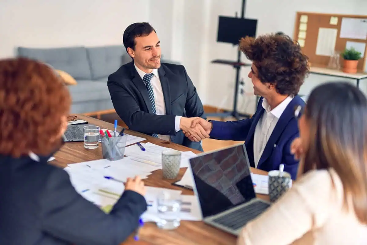 Professionals in a business meeting, shaking hands across a table with laptops and documents.