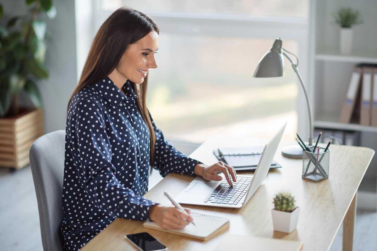 A smiling woman in a blue dotted blouse works on her laptop at a modern office desk. testimonial