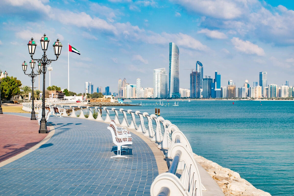 A scenic view of the Abu Dhabi skyline with a coastal walkway, street lamps, and a UAE flag.
