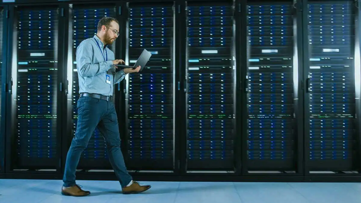 A systems engineer walks through a data center with a laptop, monitoring server racks, symbolizing distributed systems management.