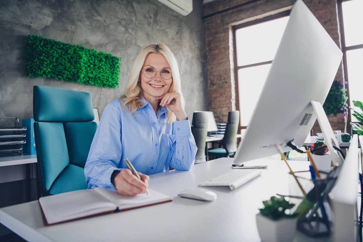Professional company secretary smiling at her desk, writing in a notebook beside a desktop computer. testimonial