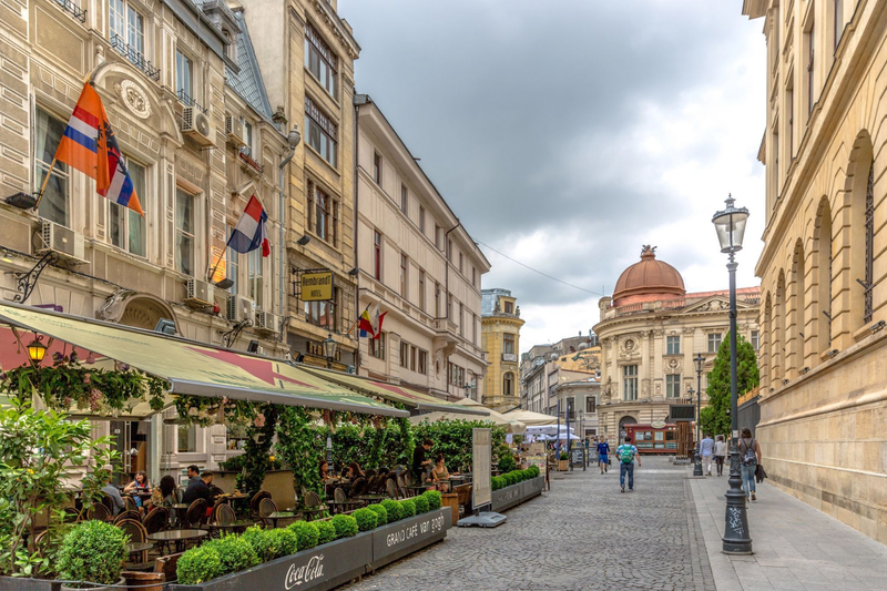 A cobblestone street in Bucharest, Romania, lined with outdoor cafés, historic buildings, and people walking under a cloudy sky.