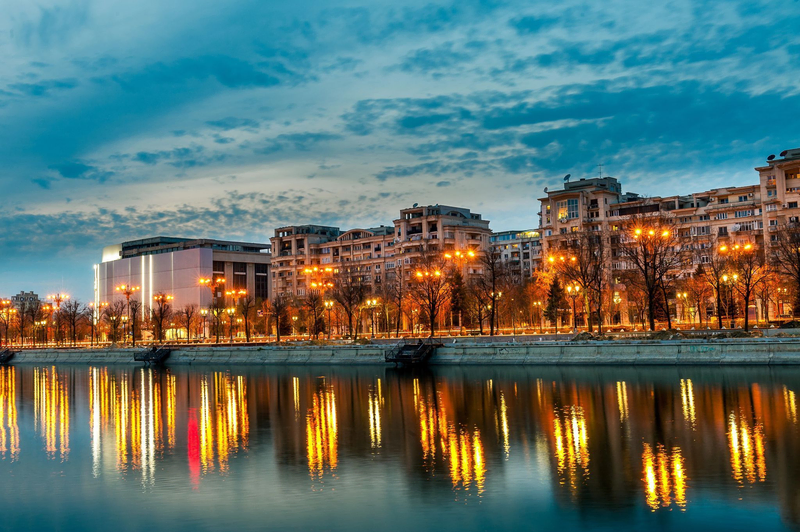 Evening view of central Bucharest along Splaiul Unirii, with city lights reflecting in the Dâmbovița River under a blue twilight sky.