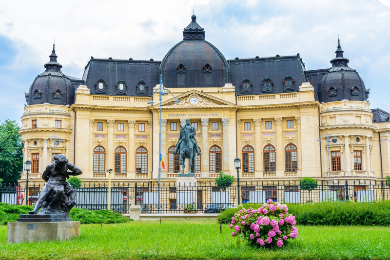 The historic Central University Library of Bucharest, Romania, with its grand façade, domed roof, and equestrian statue in front, viewed from a garden with flowers.