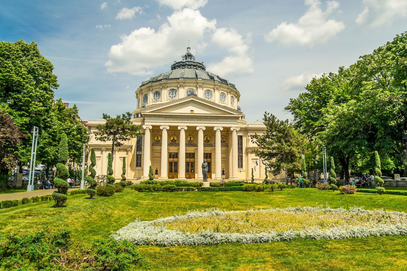 The Romanian Athenaeum in Bucharest, Romania, with its domed roof, grand columns, and surrounding gardens on a bright summer day.
