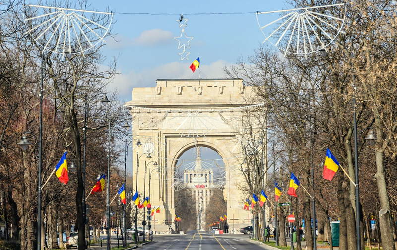 The Triumphal Arch in Bucharest, Romania, viewed from a flag-lined avenue decorated with holiday lights.